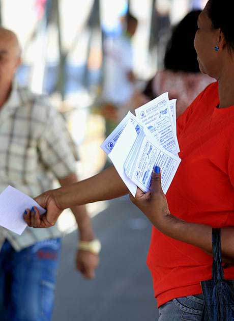 Person in a red shirt handing out flyers on a busy street while holding several leaflets in one hand.