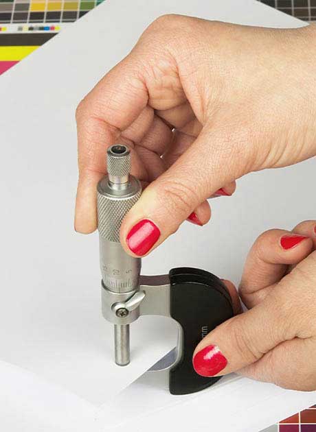 A close up view of hands with red painted nails using a metal micrometre tool to measure the thickness of a sheet of white paper on a colour testing surface.