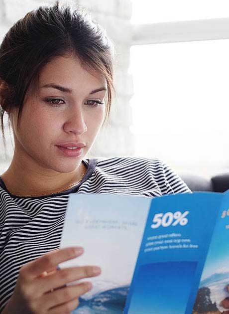 Woman wearing a striped shirt sitting indoors reading a blue promotional leaflet showing a 50 percent discount offer.